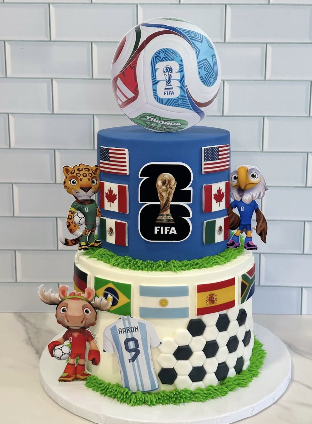Blue and white two-tier FIFA World Cup 2026 themed cake with team flags and a fondant soccer ball